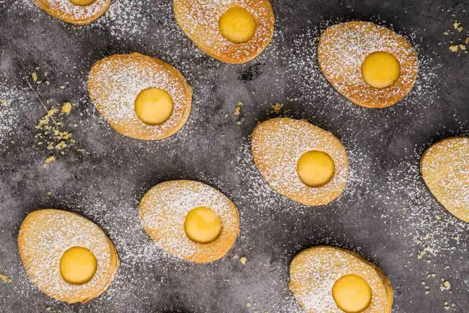 Oval-shaped cookies with powdered sugar and round cut-out centers are arranged on a dark surface, with some crumbs scattered around.