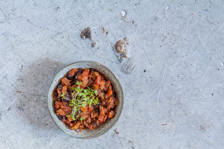 A bowl of stew garnished with microgreens sits on a textured, gray surface.