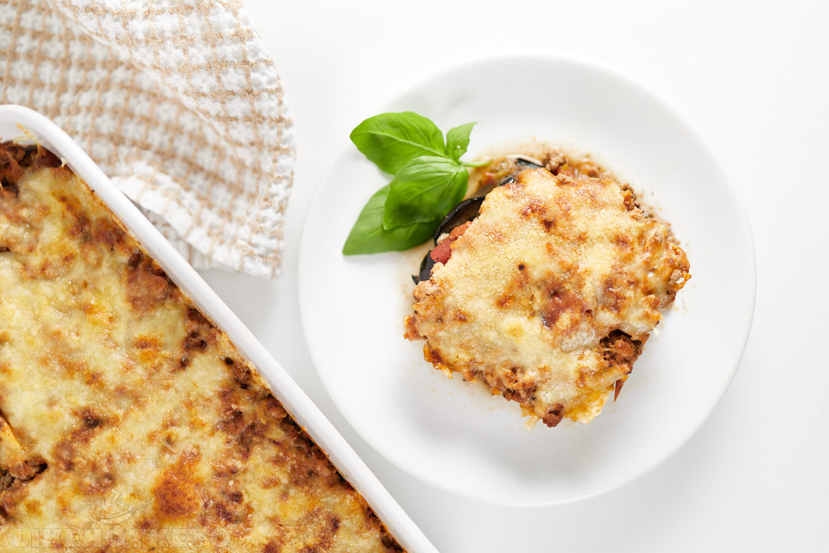 A slice of baked lasagna served on a white plate with a basil garnish, next to a casserole dish of lasagna and a checkered kitchen towel.