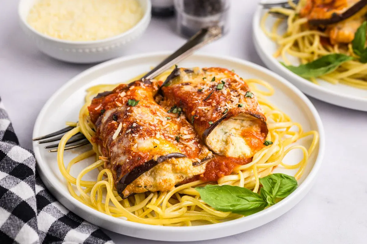 A plate of spaghetti topped with rolled eggplant slices filled with ricotta, covered in tomato sauce and garnished with basil, with a fork and knife on the plate.