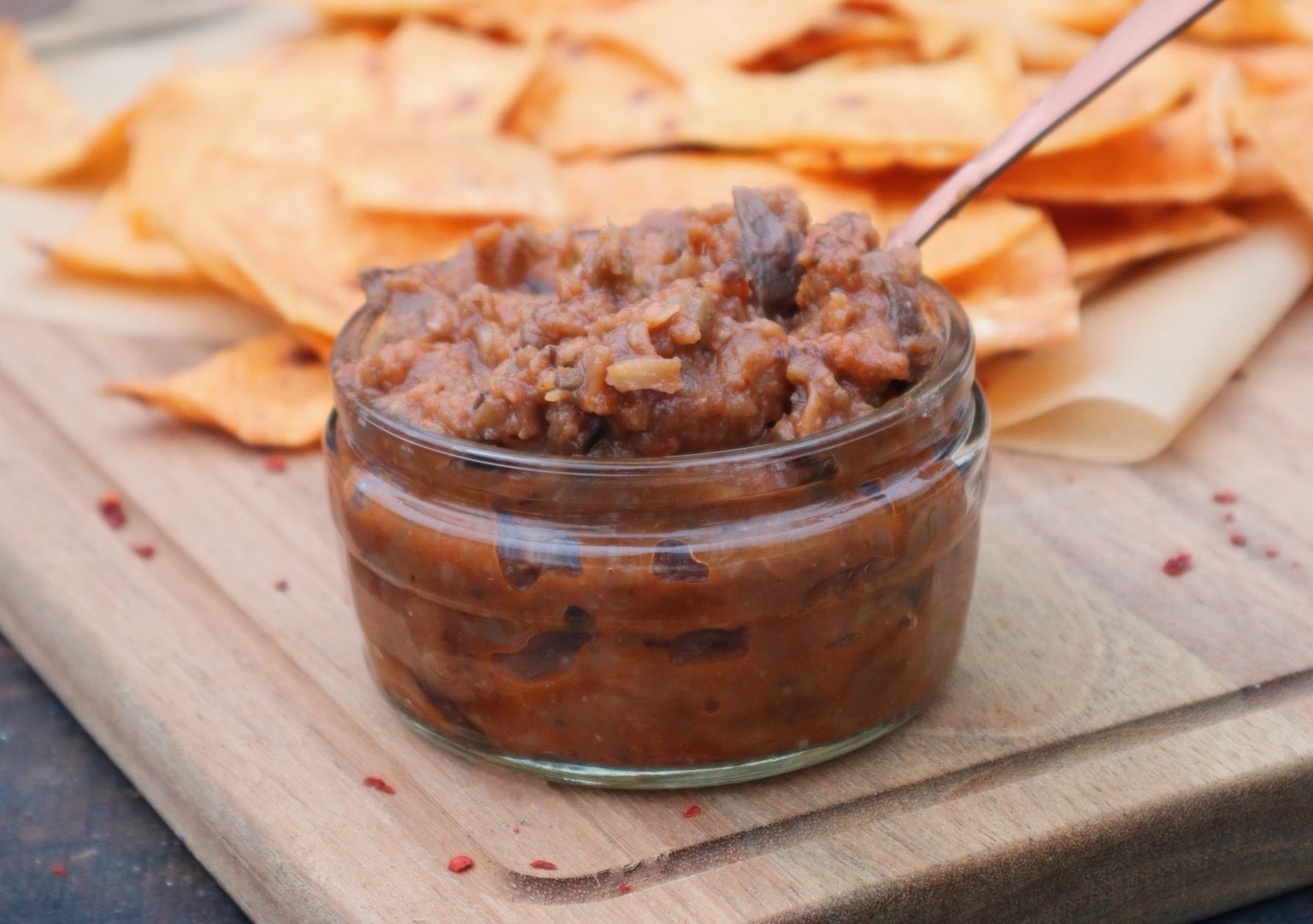 A small glass jar of refried beans with a spoon, placed on a wooden board next to a pile of tortilla chips.