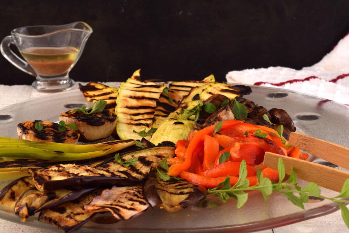 A glass plate with assorted grilled vegetables, including eggplant, bell pepper, and zucchini, garnished with fresh herbs. A small pitcher of dressing is in the background.