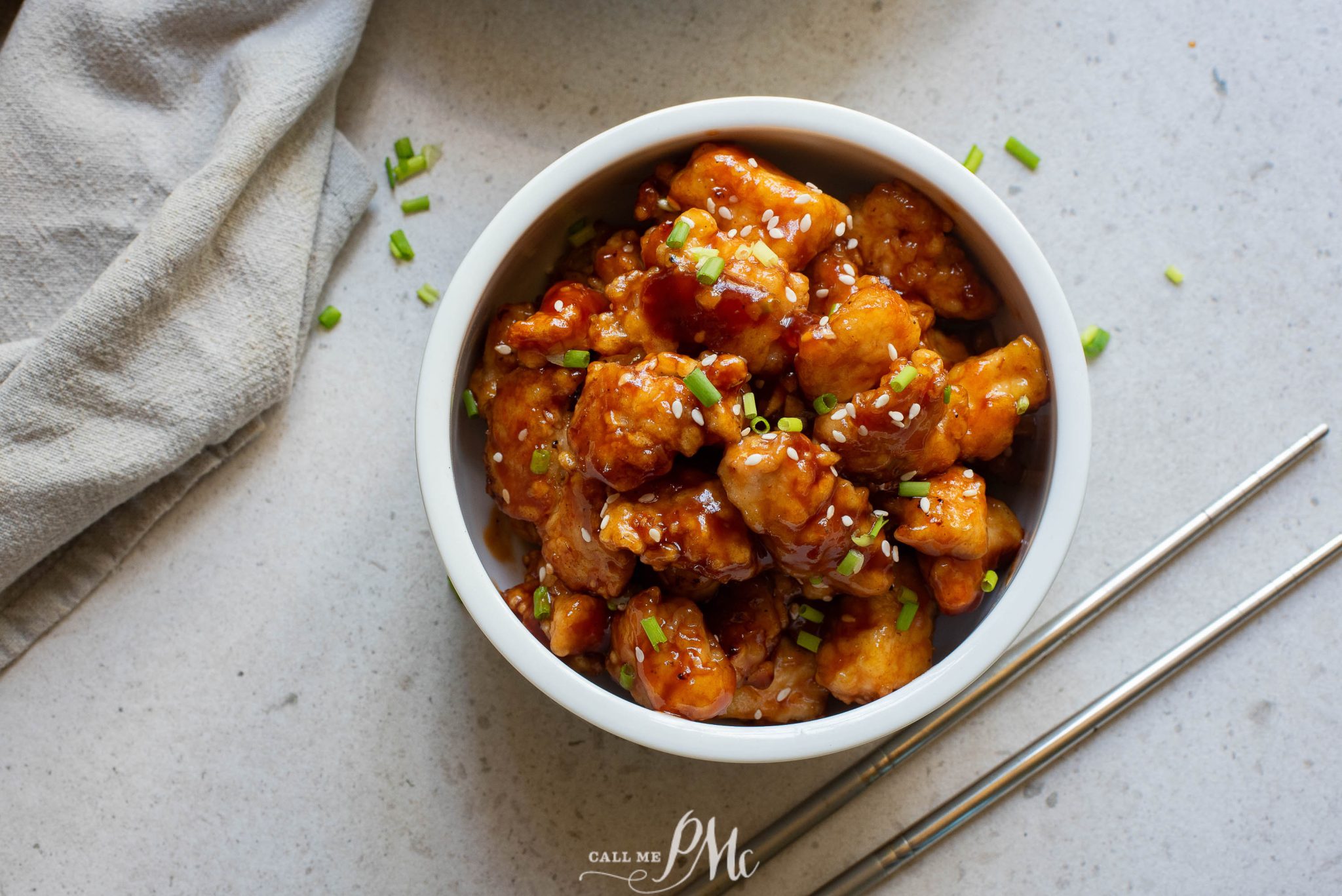 A white bowl of glazed orange chicken garnished with chopped green onions and sesame seeds, placed on a gray surface next to a pair of chopsticks and a napkin.