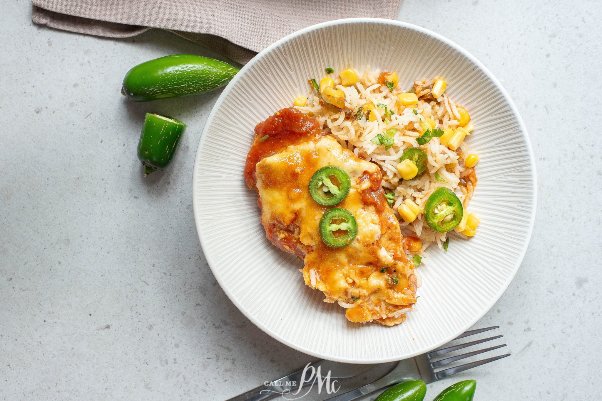 A plate with cheesy enchiladas topped with jalapeño slices, served with corn and rice. Green jalapeños and a fork are nearby on a light surface.