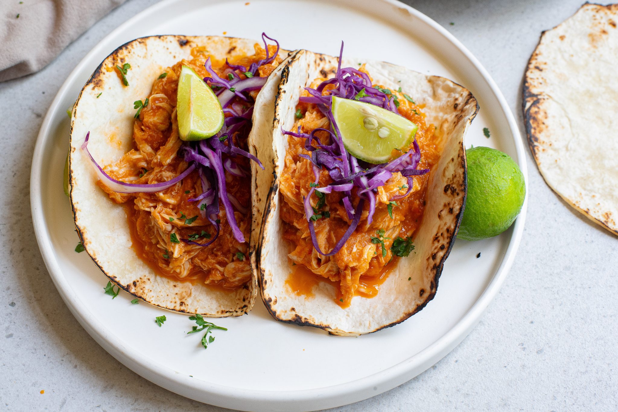 Two tacos filled with shredded chicken in a red sauce, topped with sliced red cabbage and lime wedges, served on a white plate with a lime half on the side.
