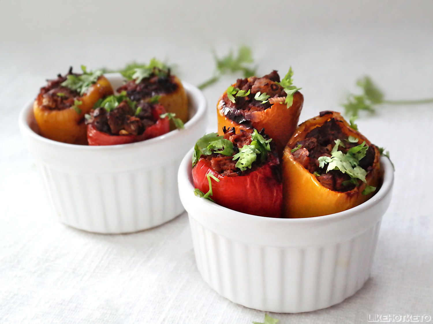 Two white ramekins filled with stuffed mini bell peppers topped with chopped herbs, placed on a white surface.