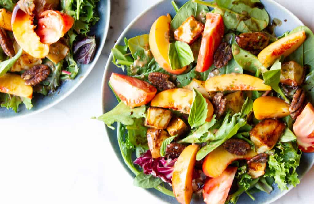 Plates of mixed green salad topped with sliced peaches, tomatoes, basil leaves, pecans, and a balsamic dressing.