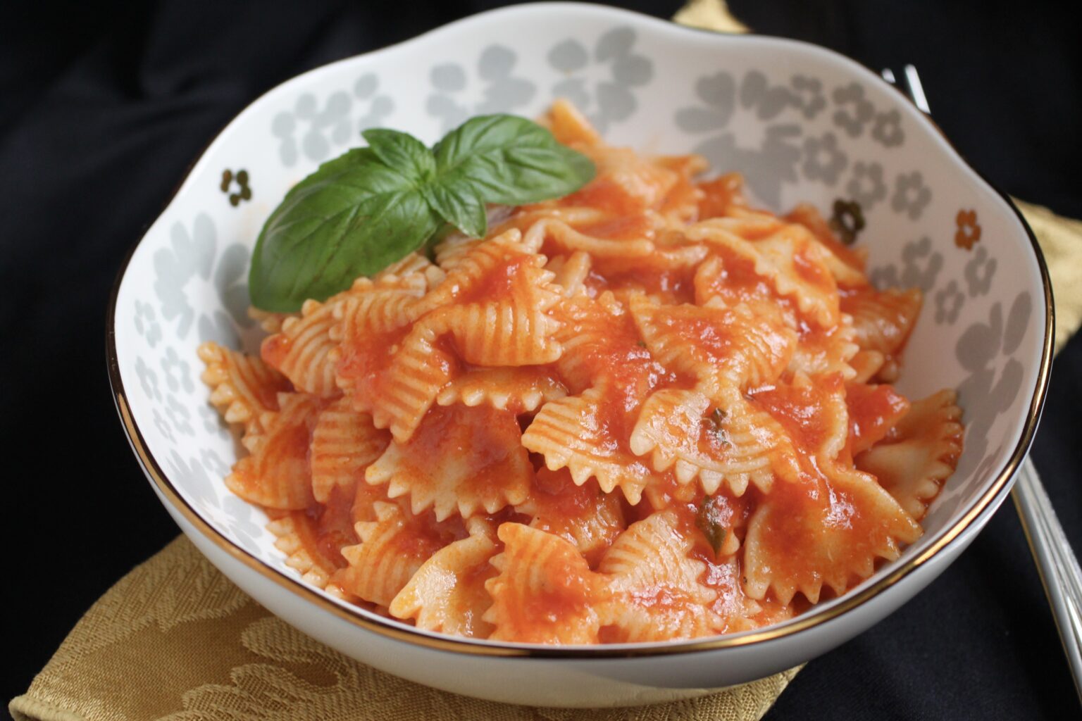 A bowl of bowtie pasta with tomato sauce, garnished with a fresh basil leaf, sits on a patterned dish against a dark background.