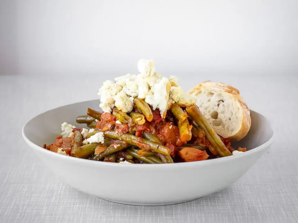 A bowl of cooked green beans in tomato sauce, topped with crumbled cheese, served with two slices of bread on the side.