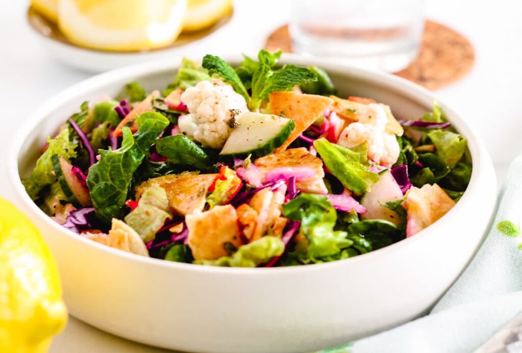 A bowl of fresh salad with lettuce, red cabbage, cauliflower, pita chips, and herbs on a white surface.