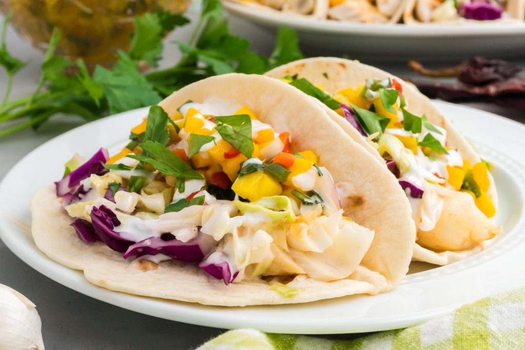 Two soft tacos filled with fish, shredded cabbage, diced mango salsa, and fresh herbs sit on a white plate.