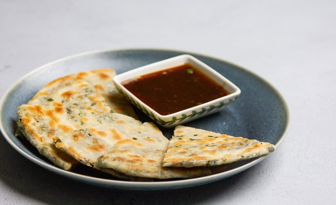 A plate with sliced scallion pancakes arranged around a square dish of dipping sauce on a gray surface.