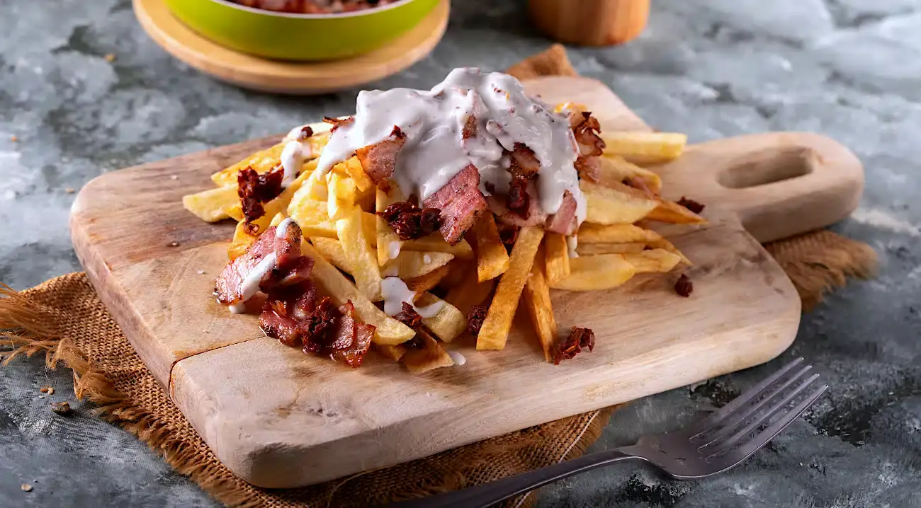 French fries topped with pieces of meat and white sauce are served on a wooden board, with a fork placed nearby.