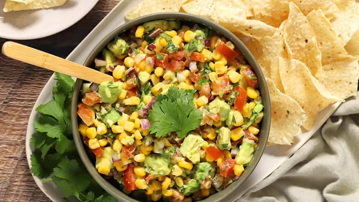 A bowl of corn, avocado, and tomato salsa garnished with cilantro, served with tortilla chips on the side.