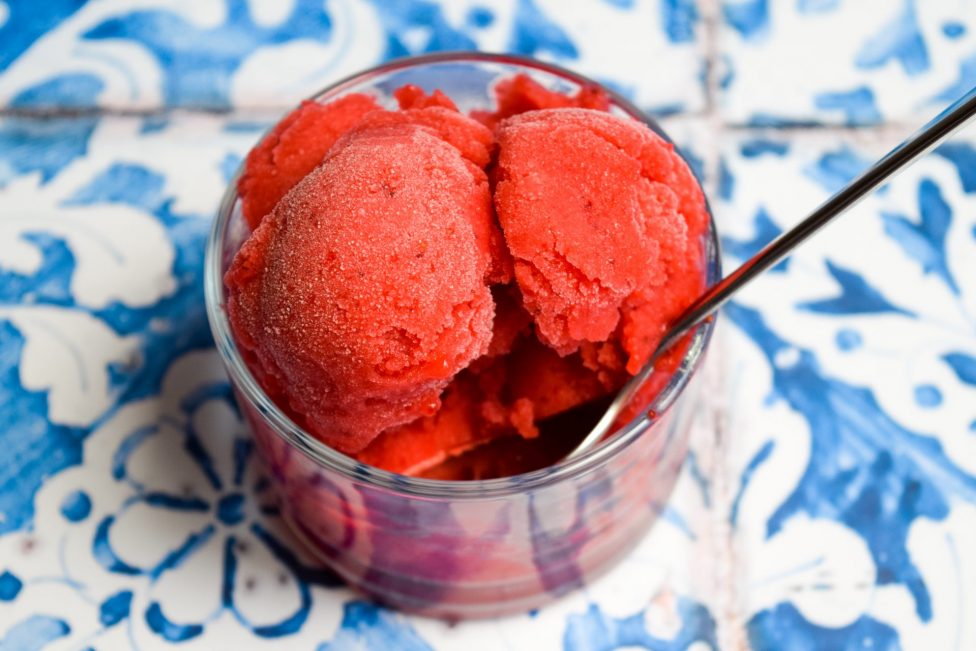 A glass bowl filled with scoops of bright red sorbet and a spoon, placed on a blue and white patterned surface.