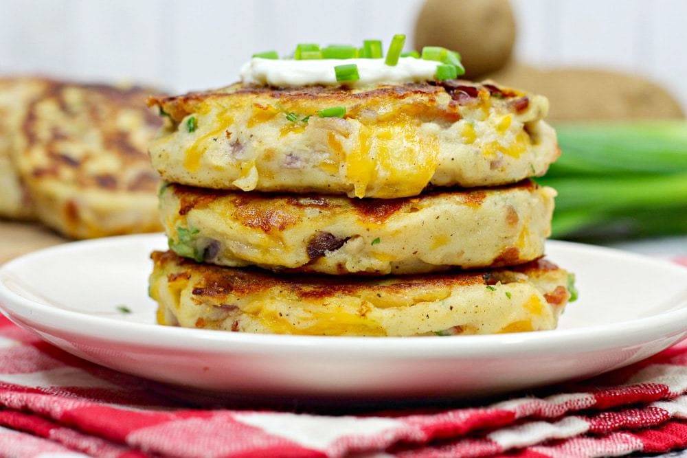 Three potato pancakes with cheese and herbs stacked on a white plate, topped with sour cream and chopped chives, on a red checked napkin.
