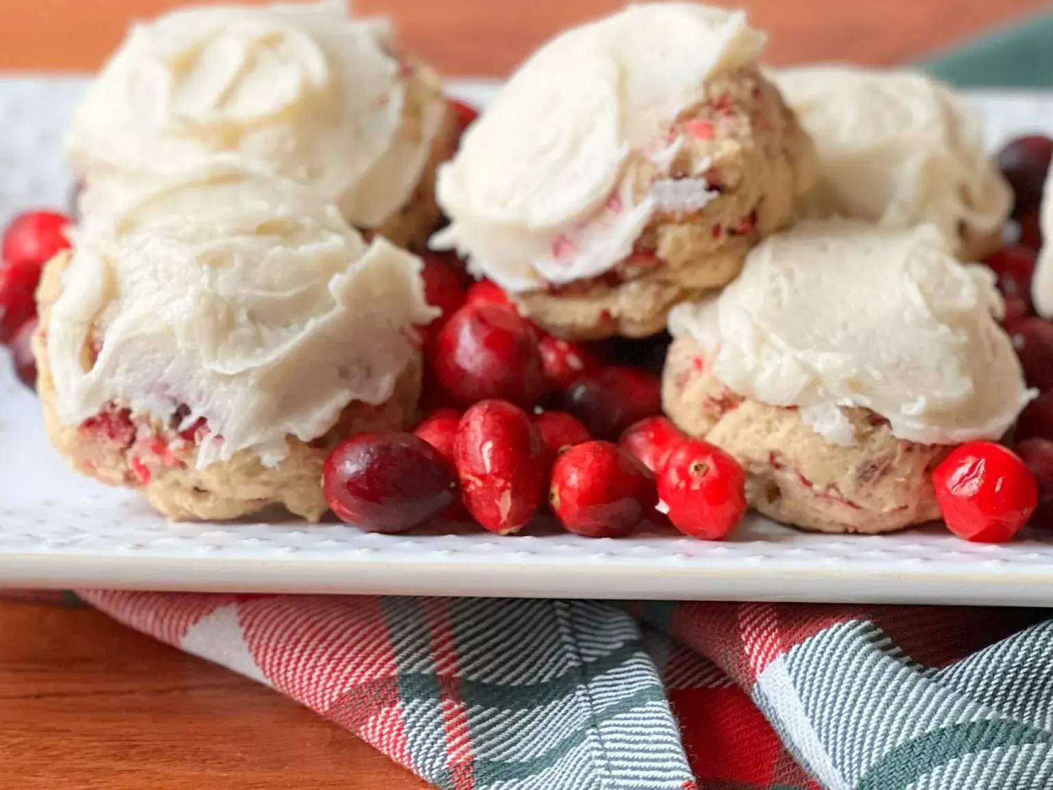 A plate of frosted cookies with visible cranberry pieces, surrounded by fresh cranberries, sits on a plaid cloth.