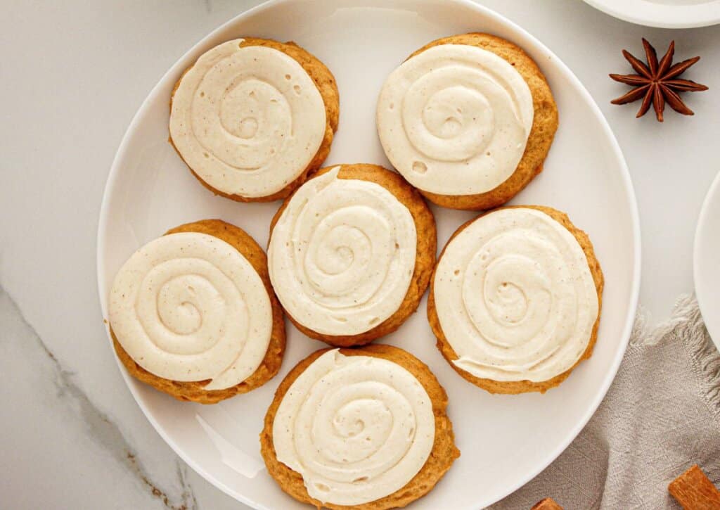 Six round cookies with swirled white icing are arranged on a white plate, viewed from above.
