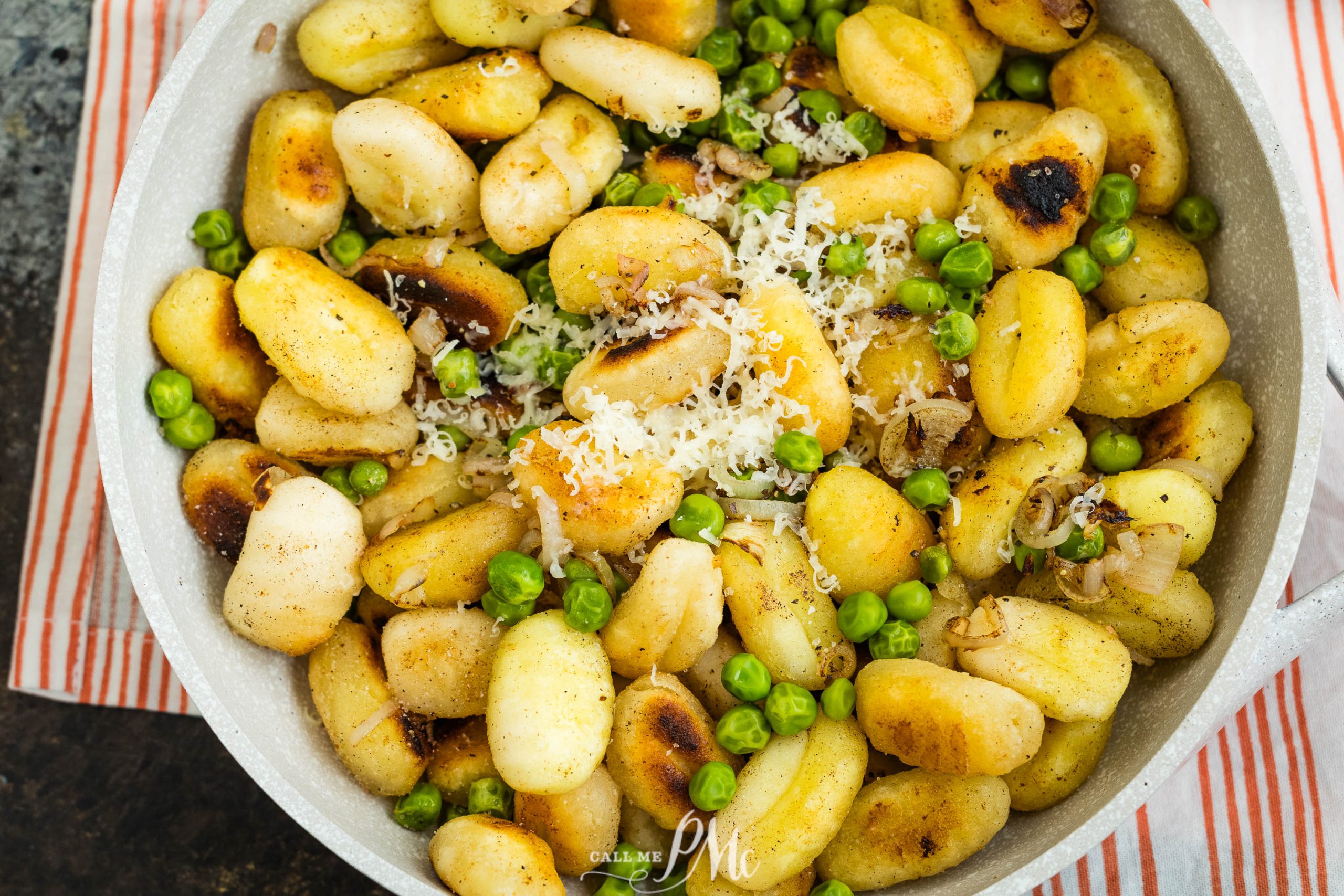 A skillet filled with golden-brown gnocchi, green peas, and grated cheese on top, set on a red-striped cloth.
