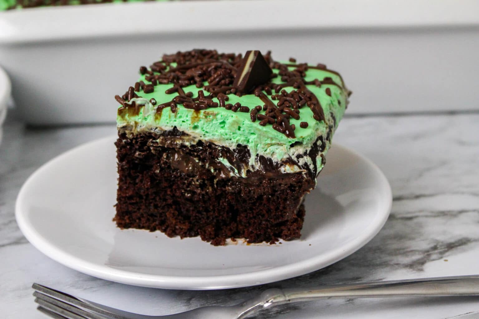 A slice of chocolate cake with green mint frosting, chocolate drizzle, sprinkles, and a chocolate mint candy on top, served on a white plate with a fork.