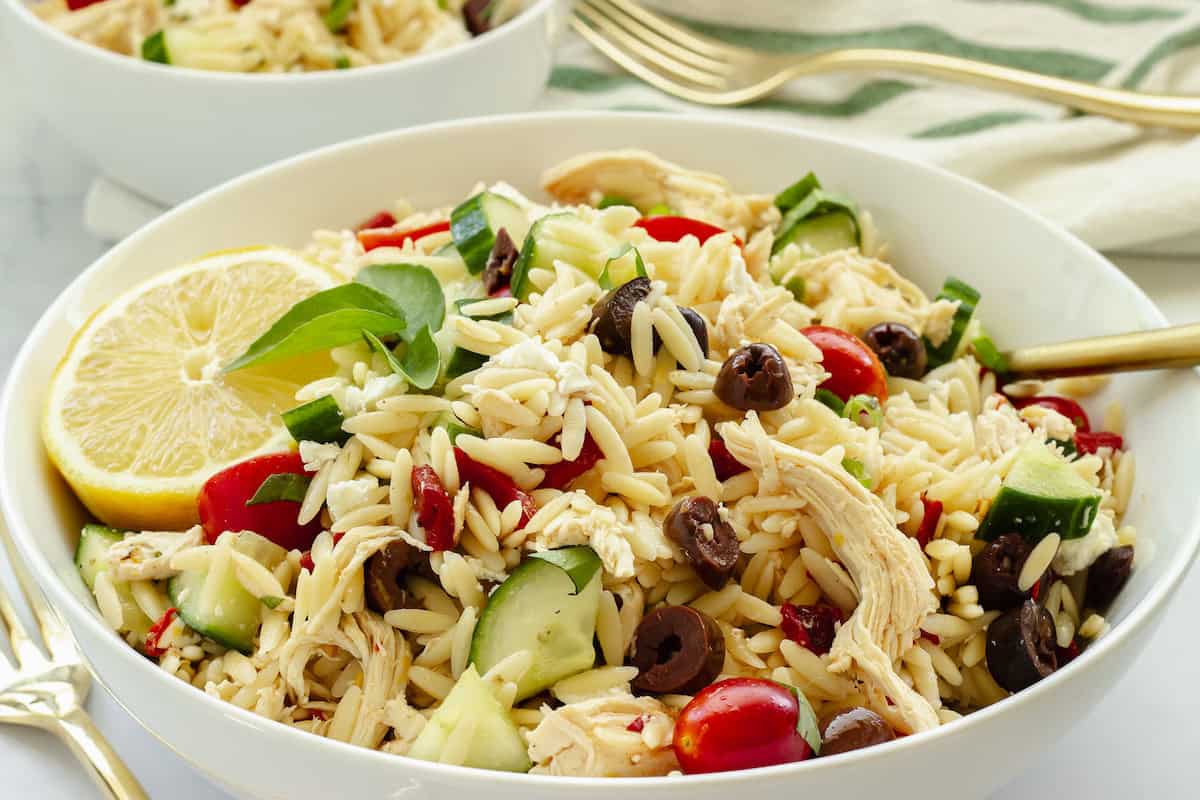 A bowl of orzo pasta salad with shredded chicken, cherry tomatoes, cucumber, black olives, feta cheese, basil, and lemon slices, with a fork and a striped napkin nearby.
