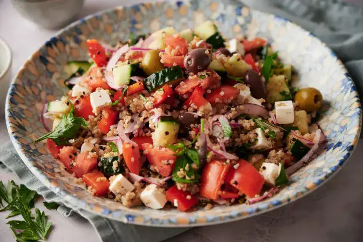 A bowl of salad with tomatoes, cucumbers, red onions, olives, feta cheese, quinoa, and greens on a patterned plate.