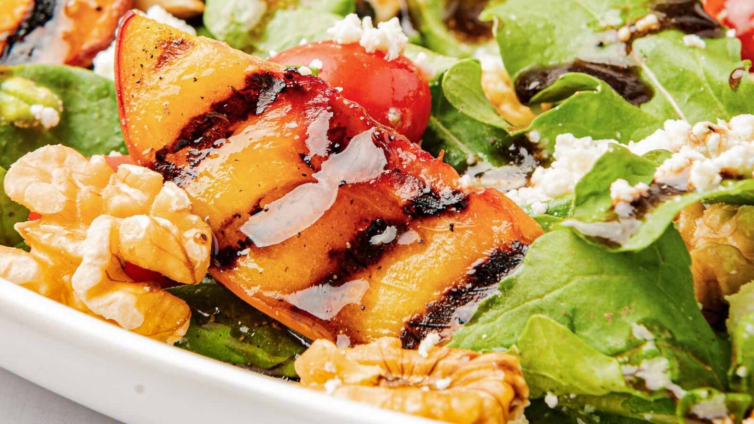 Close-up of a salad with grilled peach slices, walnuts, cherry tomatoes, mixed greens, and crumbled cheese.