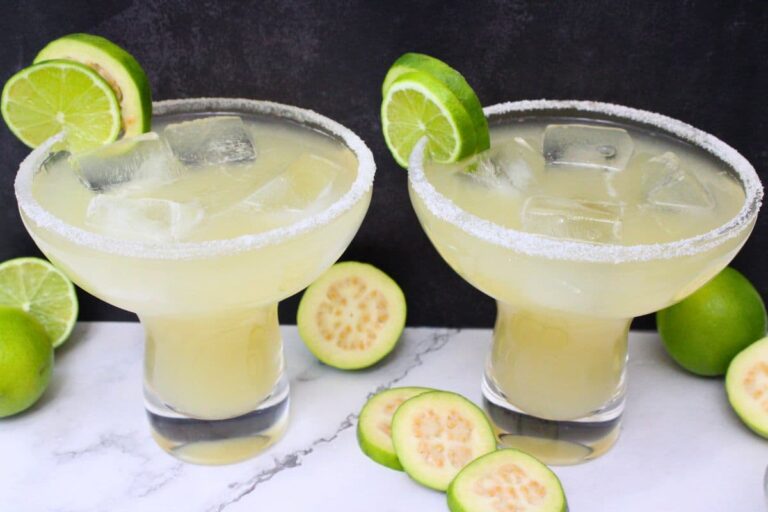 Two margarita glasses with salted rims, filled with light yellow drink and ice, each garnished with a lime slice. Sliced and whole guavas and limes are arranged around the glasses.