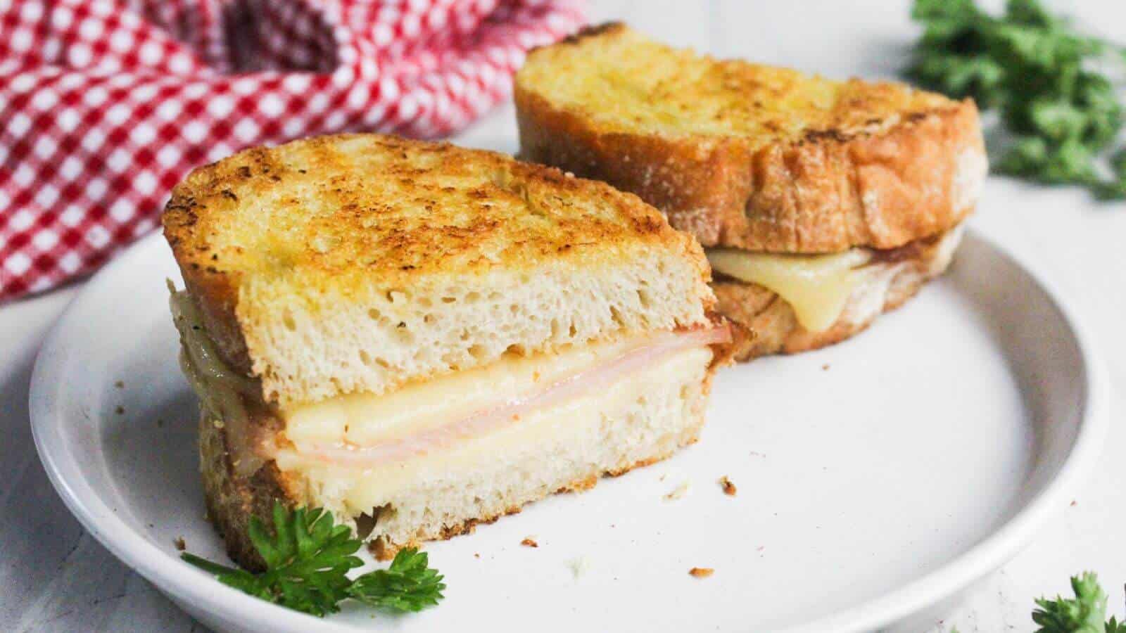 A grilled ham and cheese sandwich, cut in half, sits on a white plate with a sprig of parsley. A red and white checkered cloth is in the background.