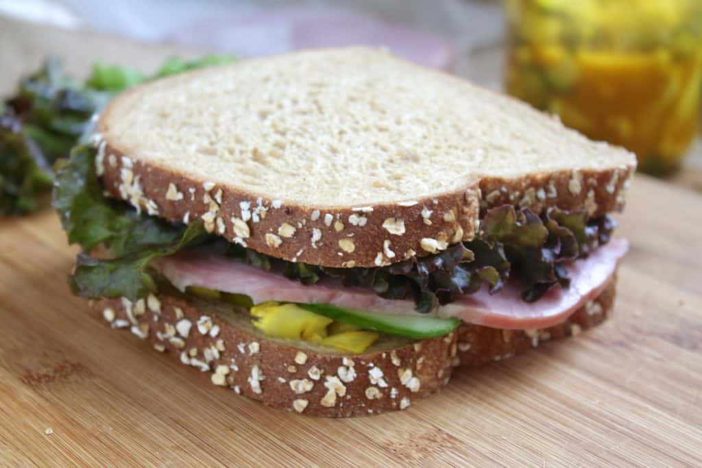 A sandwich made with oat-topped whole grain bread, lettuce, ham, sliced pickle, and avocado sits on a wooden surface.
