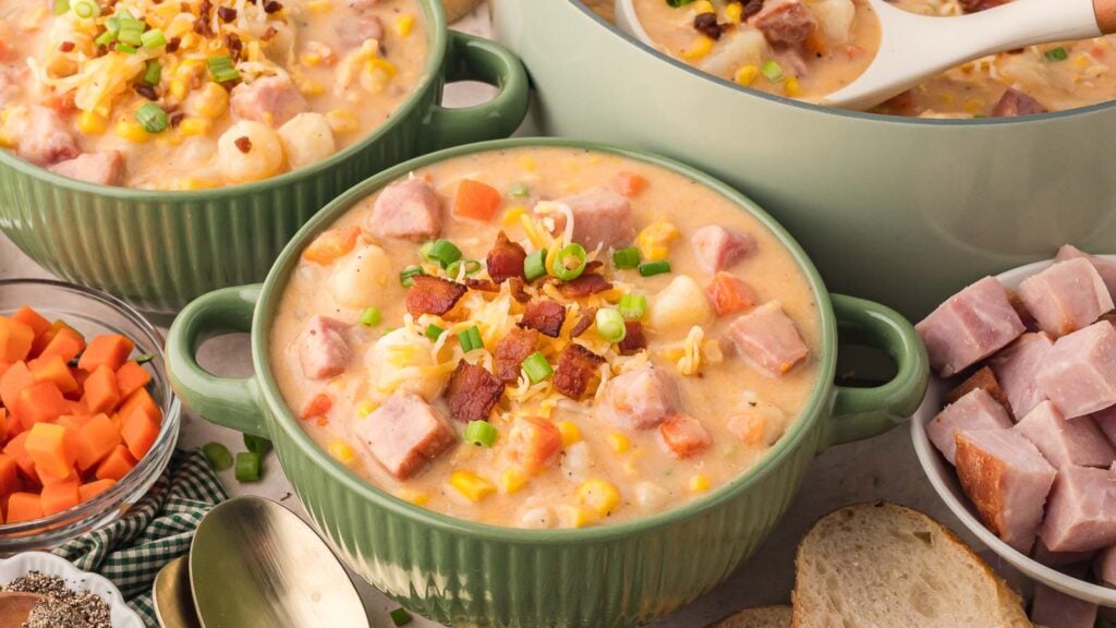 Two bowls of creamy soup with ham, corn, diced carrots, and bacon bits, garnished with green onions, next to sliced bread and soup ingredients.