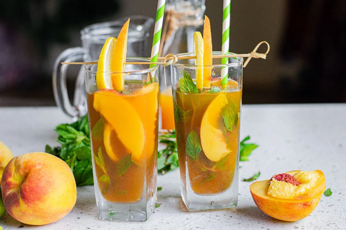 Two glasses of iced tea with peach slices, mint leaves, orange wedges, and striped straws, set on a counter with peaches and a glass pitcher in the background.