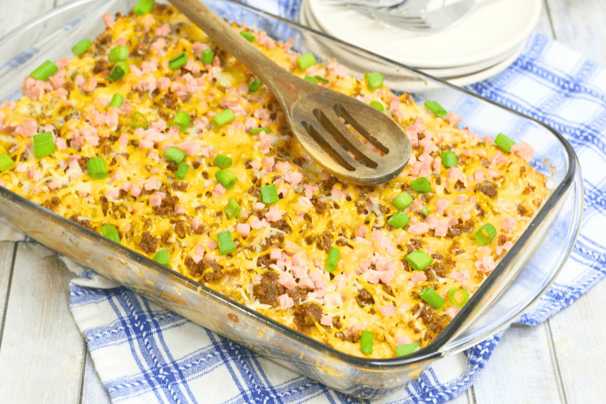 A glass baking dish holds a baked casserole topped with chopped ham, ground beef, melted cheese, and diced green onions; a wooden spoon rests on top.