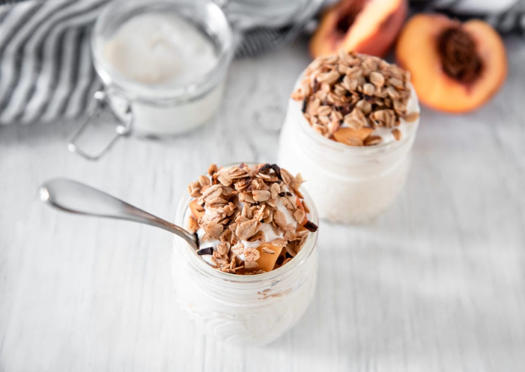 Two jars of yogurt topped with granola and peach slices sit on a white surface, with a spoon in one jar and a halved peach in the background.
