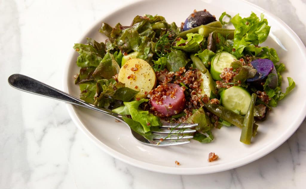 A plate of mixed green salad with sliced cucumbers, red quinoa, green beans, and assorted potatoes, with a fork resting on the plate.