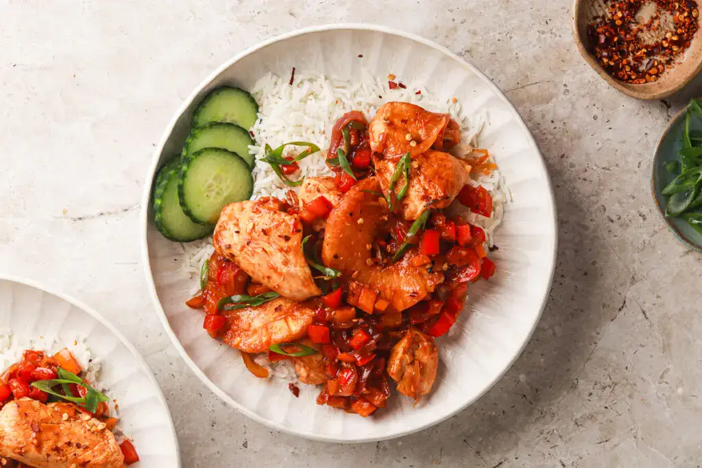 A plate of white rice topped with cooked chicken pieces, diced red bell peppers, onions, and green garnish, served with cucumber slices on the side.