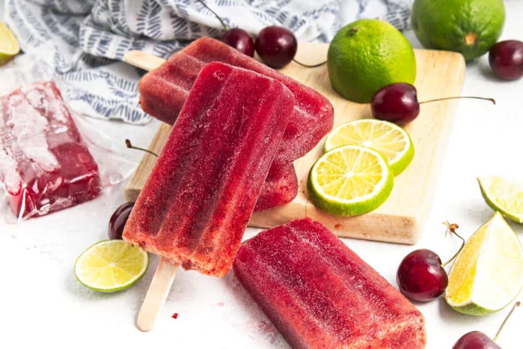 Three cherry popsicles are arranged on a white surface with fresh cherries, whole and sliced limes, a wooden board, and a patterned cloth in the background.