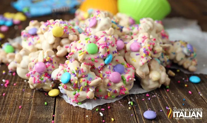 A pile of white chocolate-covered clusters topped with colorful sprinkles and candy-coated chocolates sits on parchment paper on a wooden surface.