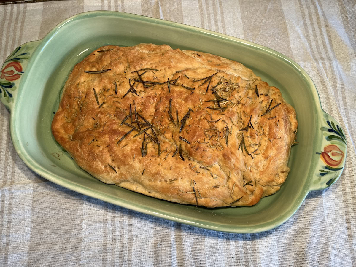 A golden-brown loaf of focaccia bread topped with rosemary sits in a green ceramic baking dish on a striped tablecloth.