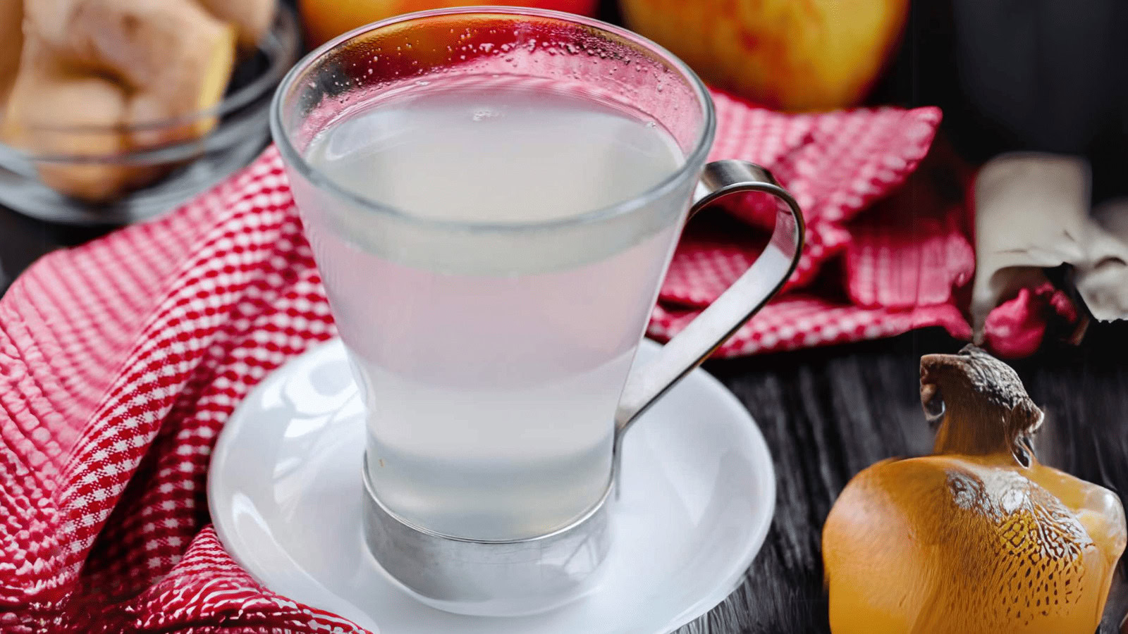 A clear glass cup filled with a cloudy liquid sits on a white saucer, surrounded by a red checkered cloth and a persimmon fruit.