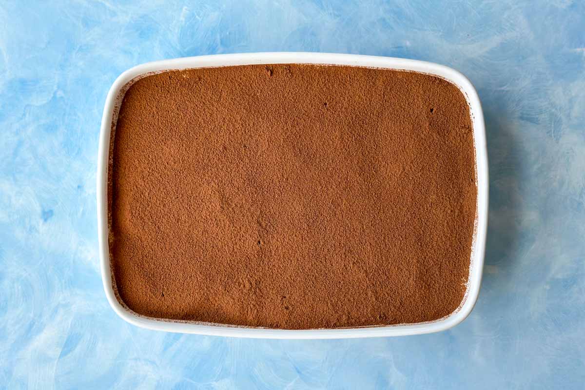 Rectangular dish filled with tiramisu, topped with an even layer of cocoa powder, placed on a light blue textured surface.