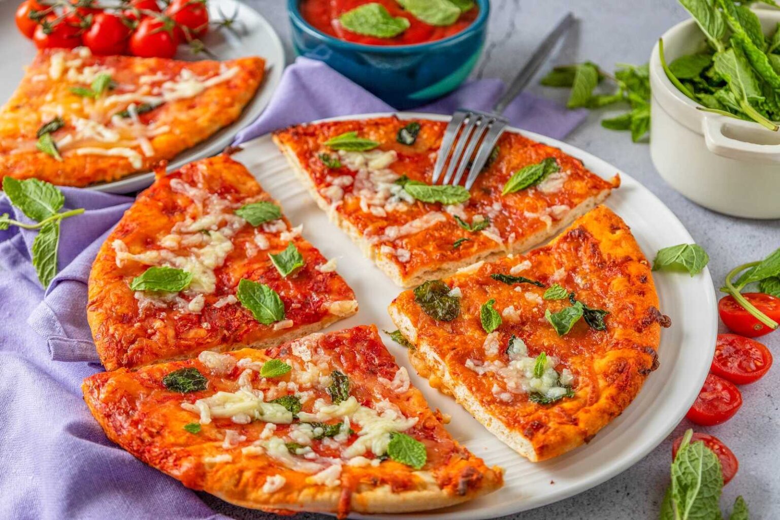 A cheese pizza topped with fresh basil leaves, sliced into six pieces on a white plate, surrounded by tomatoes, sauce, and a bowl of greens.