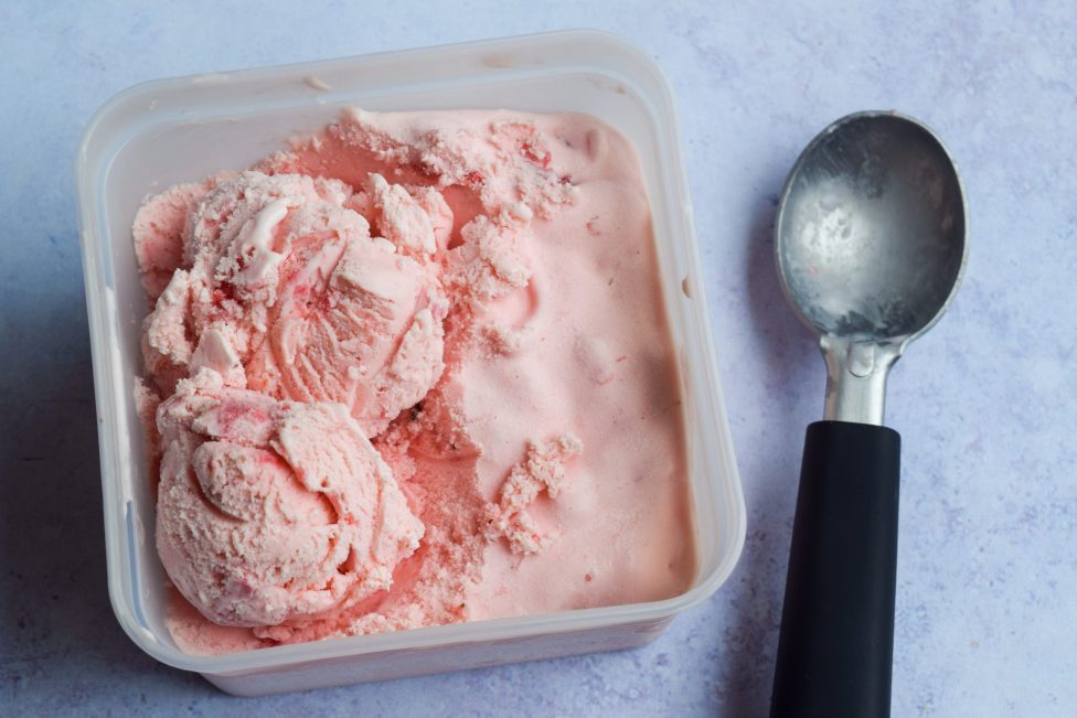 A square plastic container filled with pink ice cream sits next to a metal ice cream scooper with a black handle on a light surface.