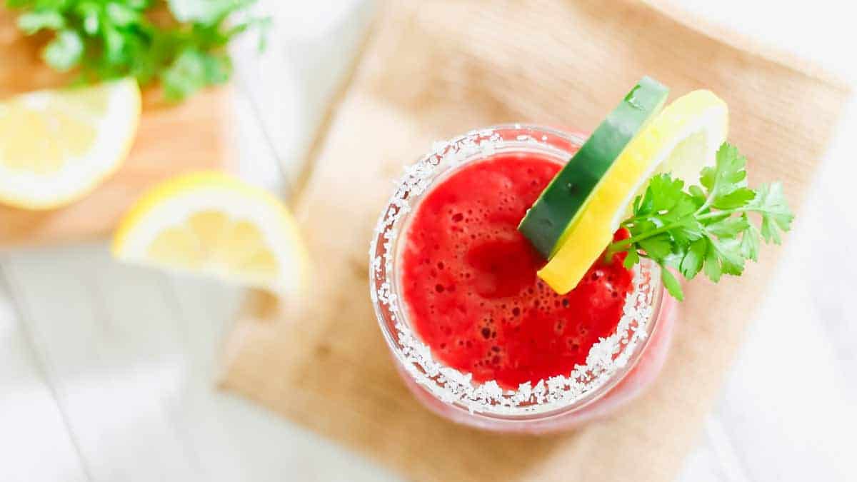 A glass of red juice with a salted rim, garnished with a cucumber slice, lemon wedge, and fresh parsley, sits on a wooden surface.