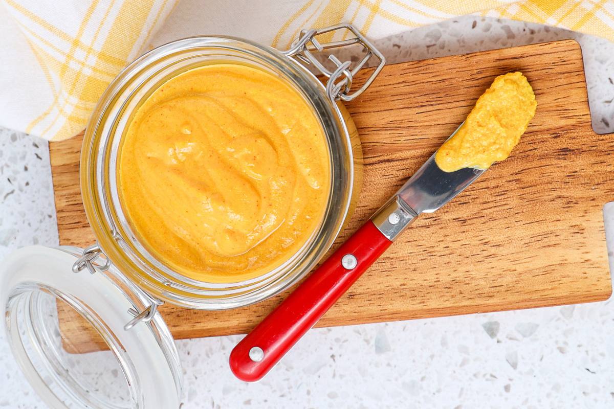 An open jar of yellow mustard sits on a wooden cutting board next to a knife with a red handle, which has mustard spread on its blade.
