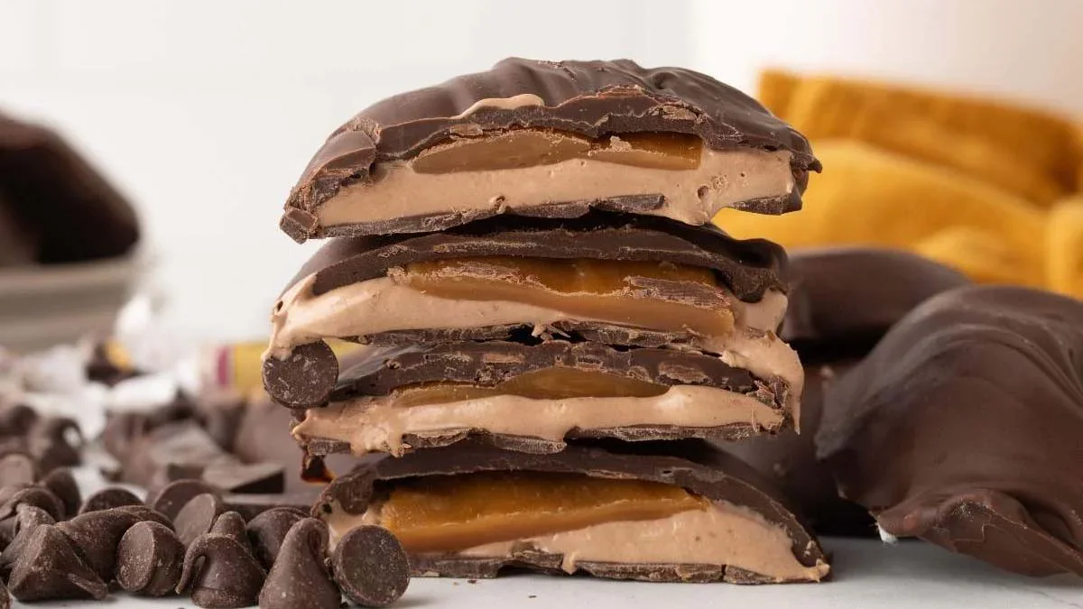 Close-up of four chocolate-covered caramel candies stacked on top of each other, with chocolate chips scattered nearby.