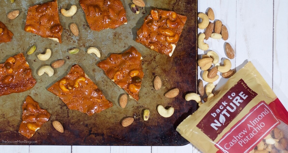 Pieces of nut brittle and scattered cashews, almonds, and pistachios on a baking sheet next to a partially open bag labeled Back to Nature Cashew Almond Pistachio.