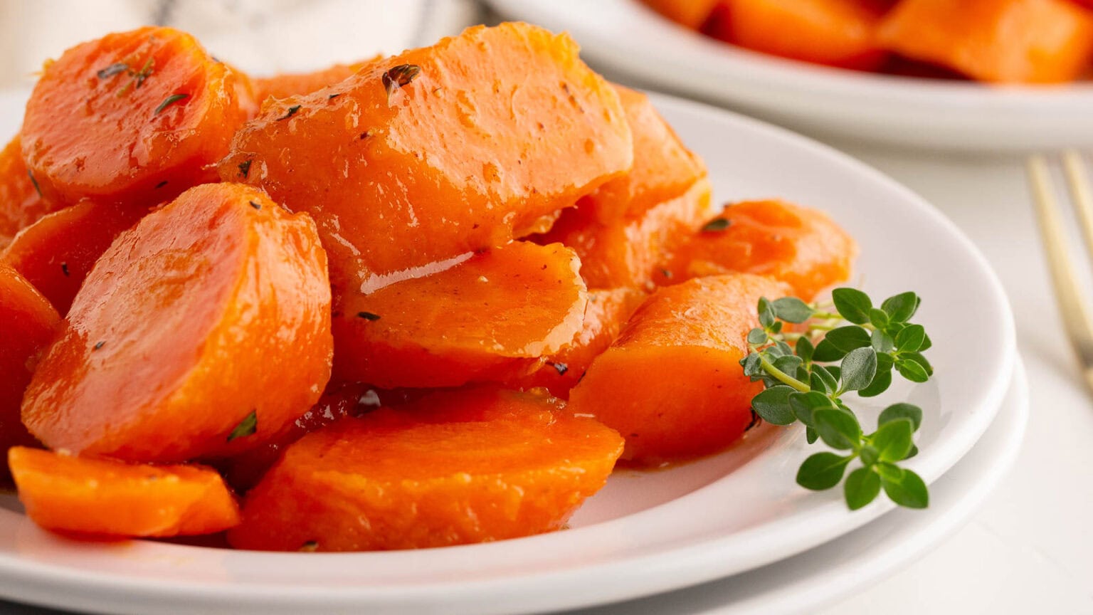 Close-up of sliced cooked carrots glazed with a shiny sauce, garnished with a small sprig of fresh herbs on a white plate.