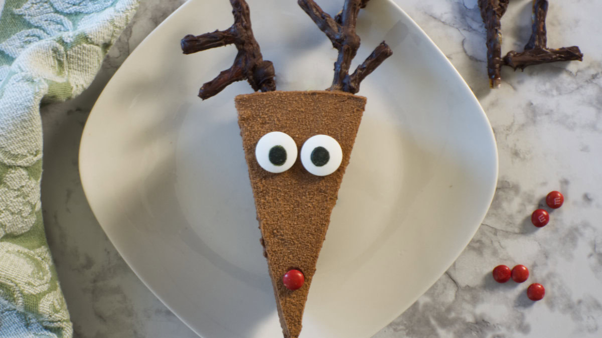 A triangular chocolate dessert decorated as a reindeer with candy eyes, a red candy nose, and chocolate antlers on a white plate.
