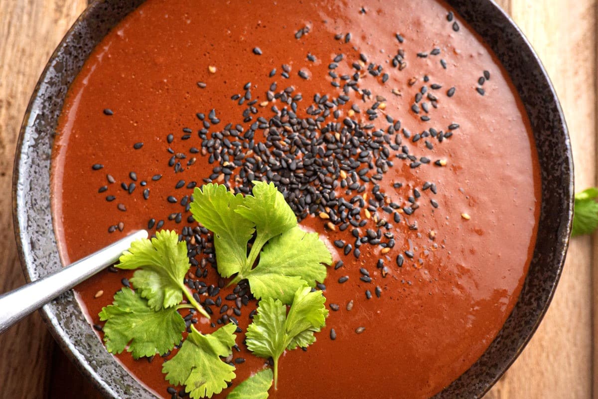 A bowl of red soup garnished with black sesame seeds and fresh cilantro leaves, with a spoon resting in the bowl.